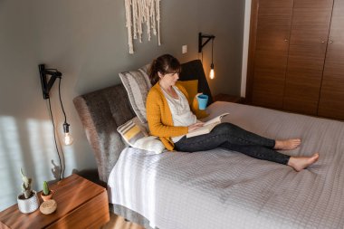 Güzel bir kadın bir fincan kahveyle yatakta kitap okuyor.