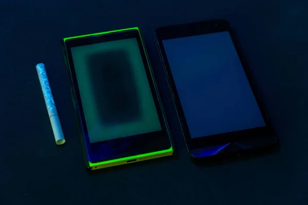 Téléphones usagés et cigarette sous lumière UV sur fond noir avec mise au point sélective — Photo