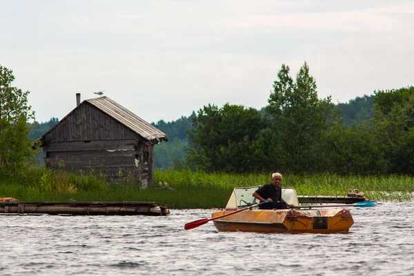 Karelia Cumhuriyeti, Rusya - 2 Temmuz 2013: Göldeki ahırın yakınında küreklerle tekneyle seyreden eski gri saçlı tabaklanmış adam. — Stok fotoğraf