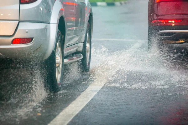 Flujo de salpicaduras de agua de lluvia de las ruedas de los coches plateados y rojos que se mueven rápidamente en la ciudad durante el día con enfoque selectivo . —  Fotos de Stock