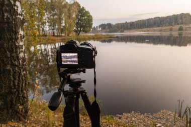 Tripod üzerindeki siyah dijital kamera, sonbahar gölü üzerindeki sisli sabah manzarasını seçici bir odak noktasıyla çekiyor.