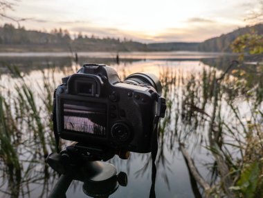 Tripod üzerindeki siyah dijital kamera, sonbahar gölü üzerindeki sisli sabah manzarasını seçici bir odak noktasıyla çekiyor.