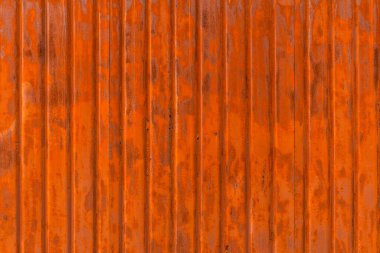 A close-up shows an old, heavily rusted corrugated metal sheet with vibrant orange-brown tones and dark corrosion marks. The vertical ridges create a strong linear texture and play of light. The aged appearance suggests long exposure to the elements.