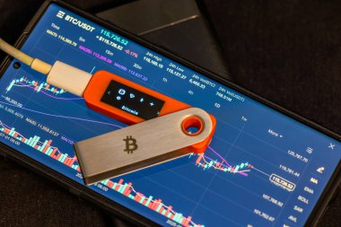 Close-up shot highlights Bitcoin hardware wallet positioned on smartphone. Screen displays detailed cryptocurrency chart, conveying serious technological atmosphere indoors.