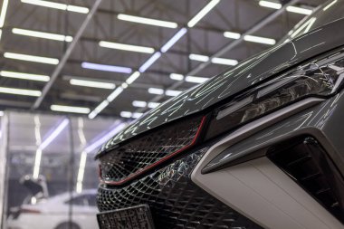 Close-up view of sleek gray car's front end highlighting headlight and grille in indoor detailing studio. Artificial lighting accentuates shiny surfaces and modern design.