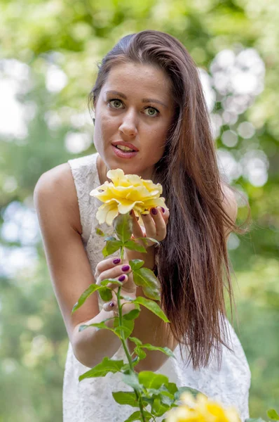 年轻女孩在一个白色礼服在夏天公园嗅到玫瑰 — 图库照片