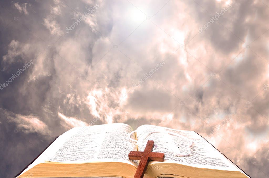 Open bible against the clouds with a wooden cross over it and bright light in the background