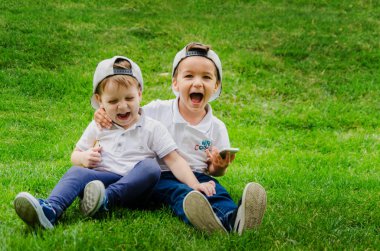 Two boys of three years in white T-shirts, jeans and caps are sitting on the grass and hugging. Two close friends, two cousins. Family relations between brothers clipart