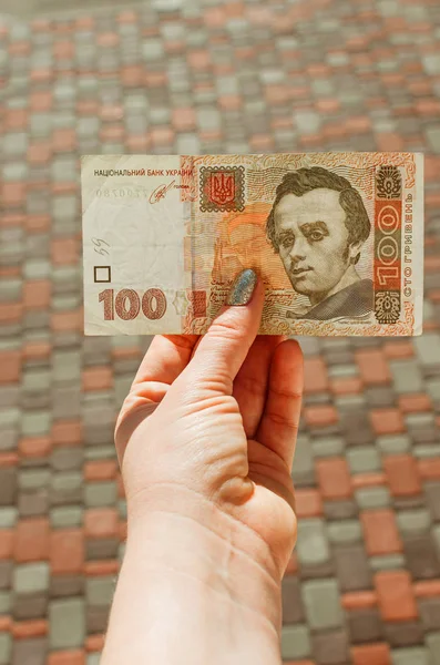 Mano femenina sostiene un billete de 100 hryvnia sobre un fondo de calle —  Fotos de Stock