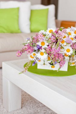 İçi çiçeklerle, ev konforu çiçeklerle dolu bir masa ve yanında da rahat bir kanepe..