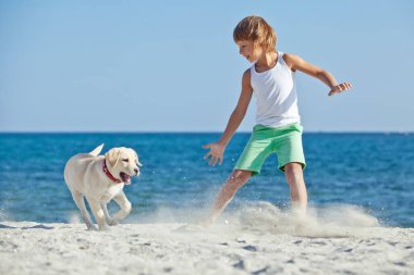 Mutlu çocuk mavi gökyüzüne karşı deniz kıyısında köpeğiyle oynuyor. En iyi arkadaşlar tatilde eğlenir..