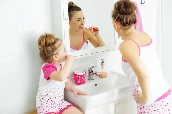 Mother and her daughter brush their teeth with toothbrushes in the bathroom at home. Mom and baby girl in home clothes. — Stock Photo, Image