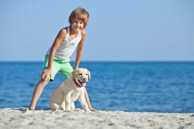 Mutlu çocuk mavi gökyüzüne karşı deniz kıyısında köpeğiyle oynuyor. En iyi arkadaşlar tatilde eğlenir..