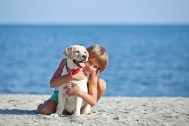 Mutlu çocuk mavi gökyüzüne karşı deniz kıyısında köpeğiyle oynuyor. En iyi arkadaşlar tatilde eğlenir..
