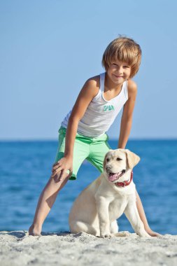 Mutlu çocuk mavi gökyüzüne karşı deniz kıyısında köpeğiyle oynuyor. En iyi arkadaşlar tatilde eğlenir, denize karşı kumsalda oynarlar..