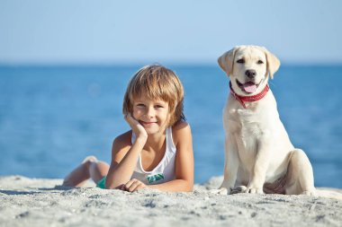 Mutlu çocuk mavi gökyüzüne karşı deniz kıyısında köpeğiyle oynuyor. En iyi arkadaşlar tatilde eğlenir, denize karşı kumsalda oynarlar..