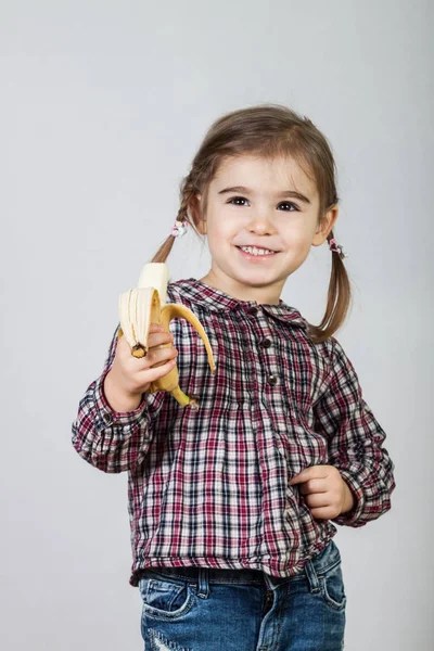 Happy little girl having fun and eating banana on gray backgroun — Stock Photo, Image