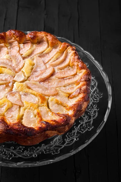 Apple pie on glass cake stand. Black wood background, high angle view.