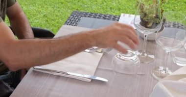 Açık hava restoranında bir masada oturan bir adam, şarap bardağını çeviriyor ve servis edilmeyi beklerken peçete alıyor..