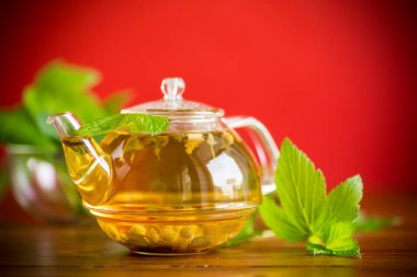 Yaz mevsiminin ferahlatıcı organik çayı, ahşap bir masanın üzerindeki cam çaydanlıkta üzümlü yapraklardan elde edilir.