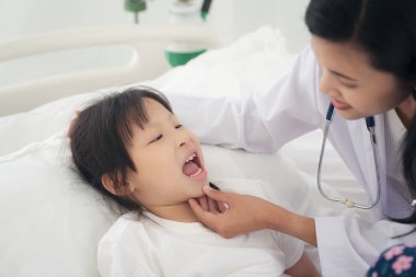 Hastanede küçük kız ağzını muayene eden kadın doktor genç bir hastanın boğazını kontrol ediyor.