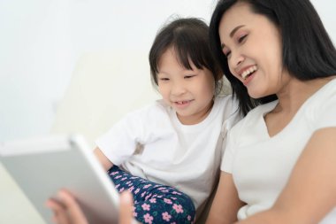 Çocuklu genç bir anne evde dijital tablet kullanıyor. Gülümseyen aile ekrana bakıyor, oyuncaklara bakıyor ya da internetten çizgi film izliyor.,