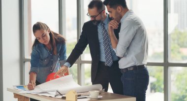 Toplantı odasında inşaat planları üzerinde çalışan çok ırklı mimarlardan oluşan bir ekip. Mühendisler ofiste proje üzerinde tartışıyorlar. Olgun iş adamı ve kadın masanın etrafında durmuş plan üzerinde çalışıyorlar..