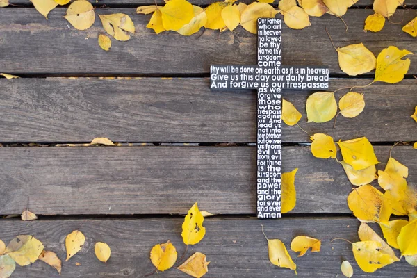 Походження осені. Білий дерев'яний хрест з молитвою Господа на потертій темній дерев'яній дошці з жовтим листям. Стокове Зображення