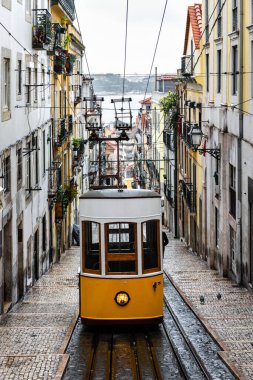 Yağmurlu bir kış gününde Lizbon 'un dar bir sokağında geleneksel sarı tramvay arka planda Tagus nehri yok olur..