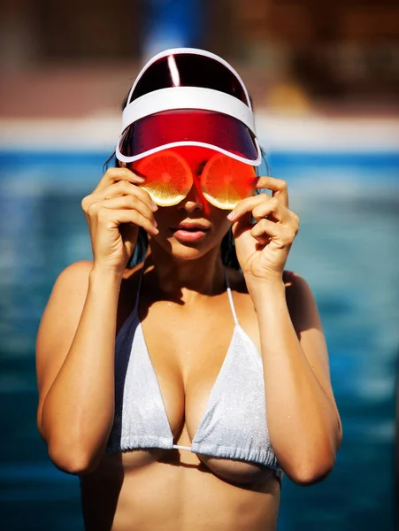 Beautiful woman in a silver swimsuit and a red sun visor with lemon slices stands in the water in the pool