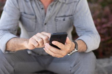 Gri elbiseli yaşlı adam sokakta oturuyor ve akıllı telefondan SMS yazıyor.