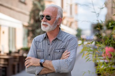 Sokakta sakallı ve güneş gözlüklü mutlu yaşlı adam poz veriyor ve gülümsüyor, portre, bulanık şehir arka planı