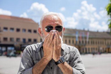 Yaşlı adam sokakta, dışarıda, sıcak yaz, alerji ve hastalık konseptinde bir mendile öksürüyor ve hapşırıyor.