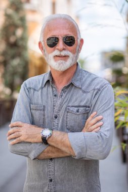 Sokakta sakallı ve güneş gözlüklü mutlu yaşlı adam poz veriyor ve gülümsüyor, portre, bulanık şehir arka planı