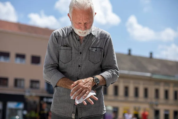 Old man cleaning hands with wet wipes in public, hot summer day