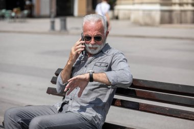 Yaşlı adam akıllı telefondan konuşuyor, saatine bakıyor, şehir içi fotoğraflarına bakıyor.