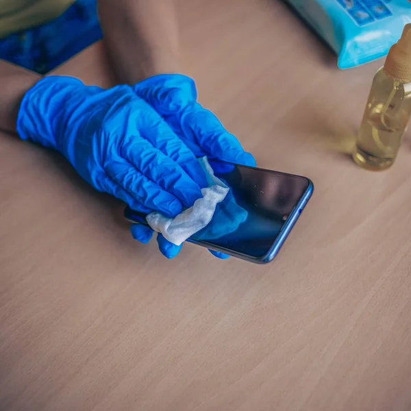 Woman's hand in blue gloves sanitizing cleaning smartphone mobile phone on wood table surface with wet wipes and alcohol