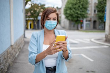 Mavi maskeli bir kadın, açık hava fotoğrafında telefonla mesajlaşıyor.