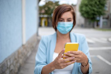 Mavi maskeli bir kadın, açık hava fotoğrafında telefonla mesajlaşıyor.