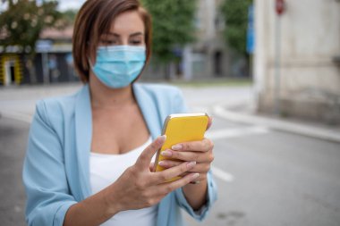 Mavi maskeli bir kadın, açık hava fotoğrafında telefonla mesajlaşıyor.