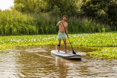 Adam paddleboarding göl kenarında ayakta. Göl kenarında su sporları yapan genç erkek. Mayo yaz tatili sırasında erkek turist.