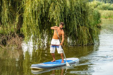 Adam paddleboarding göl kenarında ayakta. Göl kenarında su sporları yapan genç erkek. Mayo yaz tatili sırasında erkek turist.