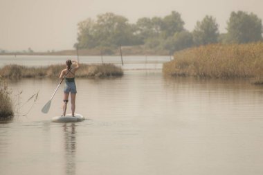 Kadın paddleboarding göl kenarında ayakta. Genç kız gölde su sporları yapıyor. Mayo yaz tatili sırasında kadın turist.