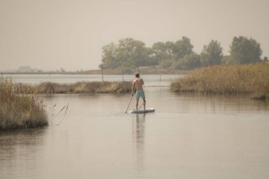 Adam paddleboarding göl kenarında ayakta. Göl kenarında su sporları yapan genç erkek. Mayo yaz tatili sırasında erkek turist.