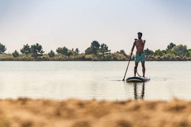 Adam paddleboarding göl kenarında ayakta. Göl kenarında su sporları yapan genç erkek. Mayo yaz tatili sırasında erkek turist.