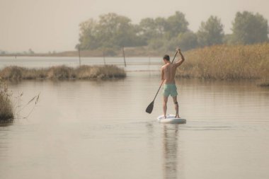 Adam paddleboarding göl kenarında ayakta. Göl kenarında su sporları yapan genç erkek. Mayo yaz tatili sırasında erkek turist.