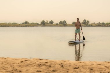 Adam paddleboarding göl kenarında ayakta. Göl kenarında su sporları yapan genç erkek. Mayo yaz tatili sırasında erkek turist.