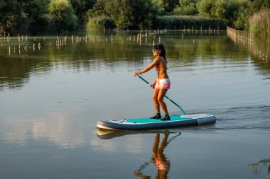 Kadın paddleboarding göl kenarında ayakta. Genç kız gölde su sporları yapıyor. Mayo yaz tatili sırasında kadın turist.
