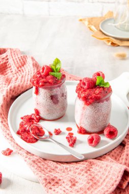 İki ayrı cam kavanoz ev yapımı ahududu chia tohumu pudingi beyaz bir tabakta sunuluyor. Sağlıklı tatlı canlı bir ahududu kompostosu ve taze ahududu ile süslenir, bir tutam nane ile süslenir. Yumuşak pembe bir havlu etrafını sarıyor.