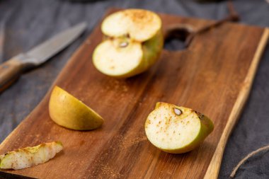 Yakın plan yatay çekimde tahta bir kesme tahtasının üzerinde dilimlenmiş yeşil elmalar görülüyor. Elma parçaları hafif tarçınlı, ve bir bıçak ve koyu bir bez yumuşak odaklı arka planda görünür.